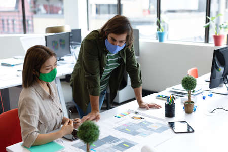 Diverse male and female colleague wearing face masks looking at blueprints and discussing. health protection hygiene in creative office during covid 19 pandemic.の写真素材