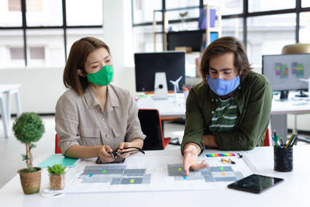 Diverse male and female colleague wearing face masks looking at blueprints and discussing. health protection hygiene in creative office during covid 19 pandemic.の写真素材