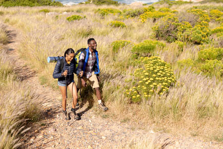 Fit afrcan american couple wearing backpacks nordic walking with poles in mountain countryside. healthy lifestyle, exercising in nature.の写真素材