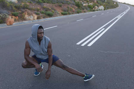 Fit african american man in sportswear stretching on a coastal road. healthy lifestyle, exercising in nature.の写真素材