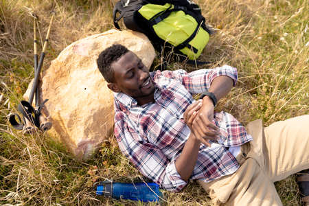 Fit african american man resting checking smartwatch and lying in mountain countryside. healthy lifestyle, exercising in nature.の写真素材