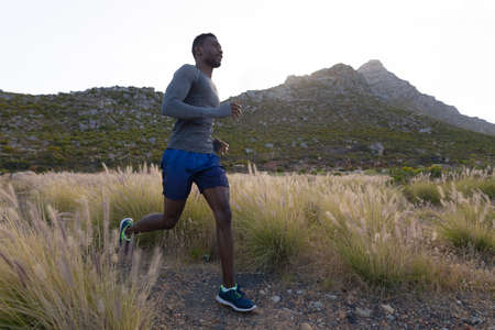 Fit african american man in sportswear running through tall grass. healthy lifestyle, exercising in nature.の写真素材