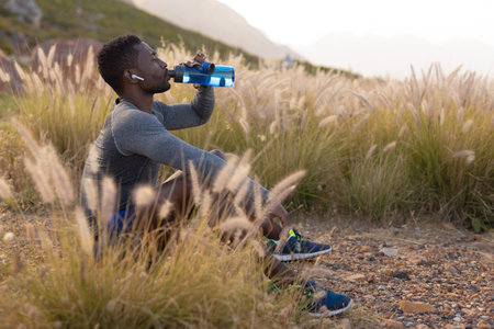 Portrait of fit african american man in sportswear resting drinking water in tall grass. listening to music with earphones in. healthy lifestyle, exercising in nature.の写真素材
