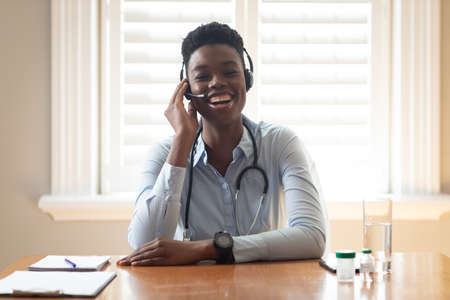 African american doctor wearing phone headset talking while having a videocall at home. distant communication and telemedicine consultation concept.の写真素材