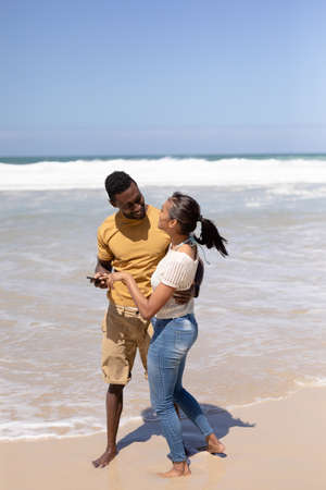 African american couple using smartphone on a beach by the sea. healthy lifestyle, leisure in...
