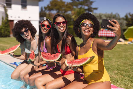 Diverse group of female friends taking selfie with watermelon sitting at the poolside. Hanging out and relaxing outdoors in summer.の写真素材