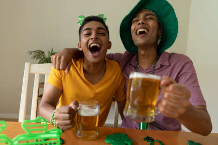 Laughing mixed race gay male couple wearing st patrick's day costumes and holding glasses of...