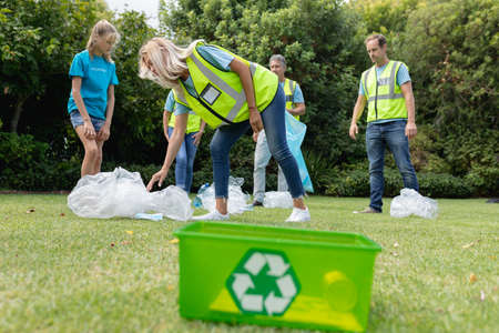 Caucasian group of men and women collecting rubbish in field with recycling box in foreground....