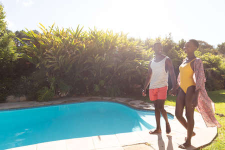 African american couple smiling at the poolside and holding hands on sunny garden terrace. staying at home in isolation during quarantine lockdown.の写真素材