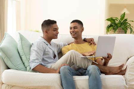 Diverse gay male couple sitting on sofa using laptop computer and embracing. staying at home in...