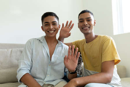 Portrait of diverse gay male couple looking at camera waving and smiling. staying at home in...