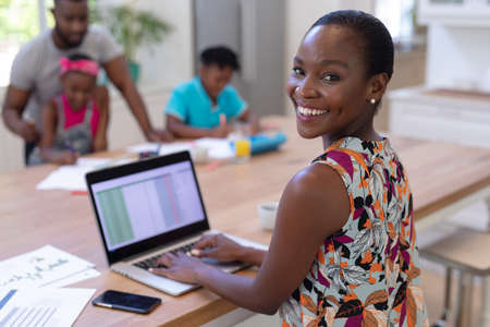 Portrait of smiling african american mother working at home using laptop with family in background. staying at home in isolation during quarantine lockdown.の写真素材
