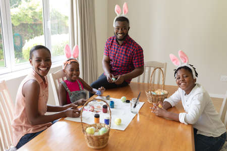 Happy african american parents with son and daughter wearing bunny ears painting easter eggs. celebrating easter at home in isolation during quarantine lockdown.の写真素材