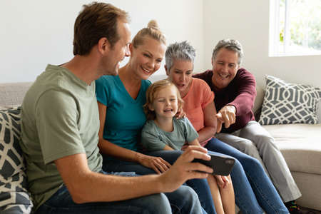 Caucasian father sitting on couch with with his wife, parents and son looking at his smartphone....