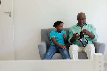 Senior african american grandfather holding vr headset sitting on couch beside grandson. happy...