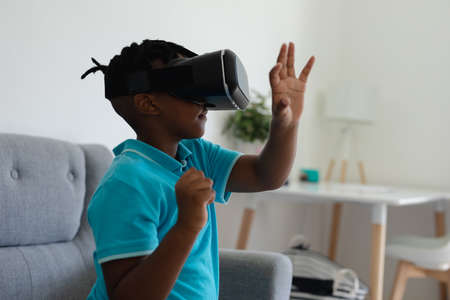 African american boy wearing vr headset sitting on couch and raising his hand. staying at home in isolation during quarantine lockdown.の写真素材