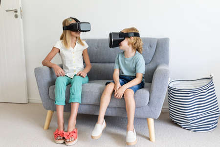 Caucasian brother and sister wearing vr headsets sitting on couch facing each other. staying at home...
