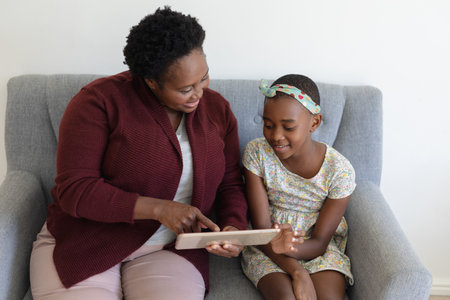 Senior african american grandmother sitting on couch beside smiling granddaughter using tablet....