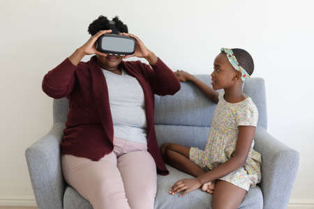 Senior african american grandmother sitting on couch wearing vr headset beside smiling...
