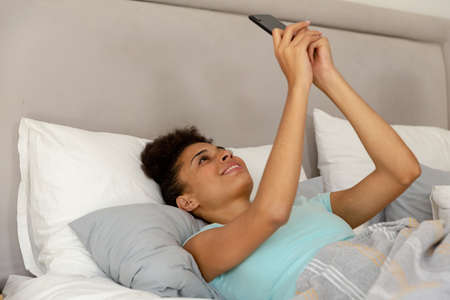 Mixed race woman lying in a bed using a smartphone and smiling. staying at home in isolation during...
