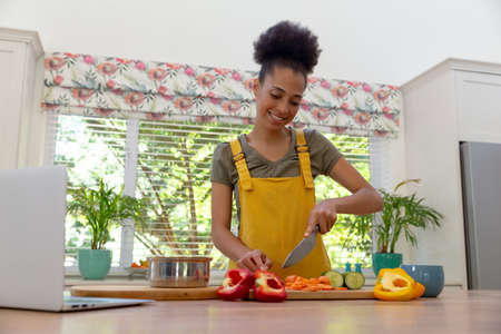 Mixed race woman cutting vegetables using laptop and smiling in a kitchen. staying at home in...