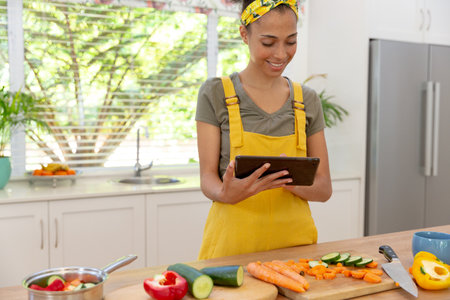 Mixed race woman cutting vegetables using digital tablet and smiling in a kitchen. staying at home...