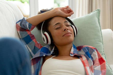 Mixed race woman lying on couch listening to music with headphones at home. staying at home in...