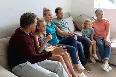 Caucasian parents, grandparents and grandchildren sitting on couch watching tv and eating popcorn....