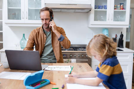Caucasian father working at home using smartphone and laptop sitting with son doing school work....