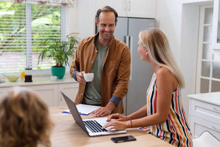 Caucasian mother working at home on laptop talking to husband, with son sitting at table. staying at...