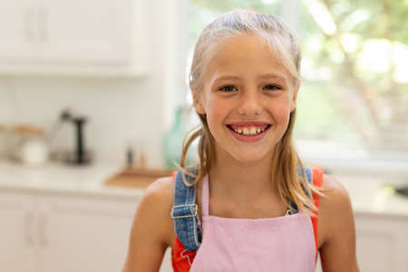 Portrait of smiling caucasian girl wearing apron standing in kitchen. staying at home in isolation...