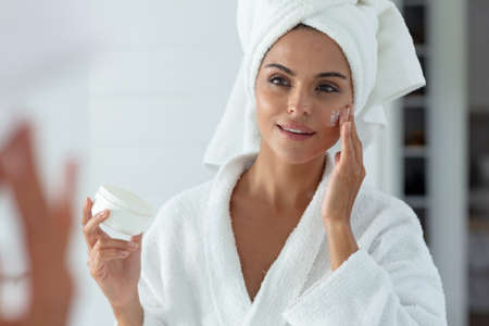 Caucasian woman wearing bathrobe standing in bathroom putting cream on face and smiling. staying at home in isolation during quarantine lockdown.の写真素材