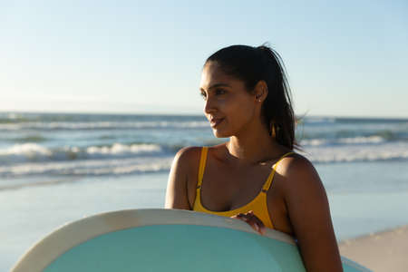 Happy mixed race woman carrying surfboard on the beach. healthy outdoor leisure time by the sea.