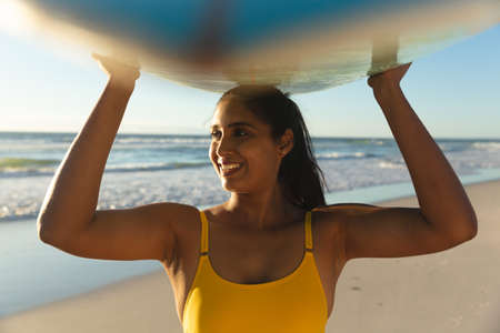 Happy mixed race woman on the beach carrying surfboard on head. healthy outdoor leisure time by the...