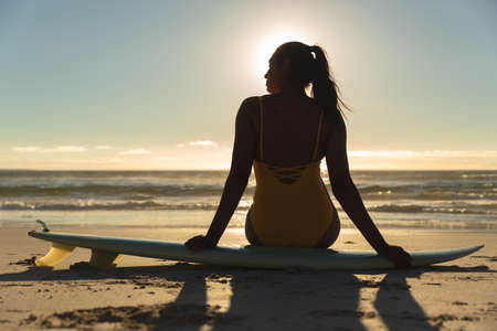 Mixed race woman sitting on surfboard on the beach. healthy outdoor leisure time by the sea.