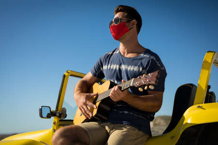 Happy caucasian man wearing face mask sitting on beach buggy playing guitar. beach stop off on...