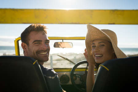 Happy caucasian couple sitting in beach buggy by the sea looking to camera. beach break on summer...