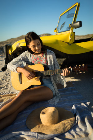 Happy caucasian woman leaning against beach buggy by the sea playing guitar. beach break on summer...