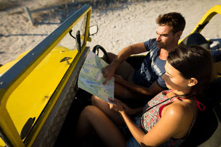Happy caucasian couple sitting in beach buggy by the sea reading roadmap. beach break on summer...