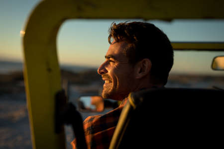 Happy caucasian man sitting in beach buggy by the sea during sunset smiling. beach break on summer...