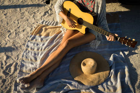 Low section of caucasian woman sitting on beach by the sea playing guitar. healthy outdoor leisure time by the sea.の写真素材