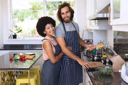 Portrait of happy diverse couple in kitchen preparing food and embracing. staying at home in isolation during quarantine lockdown.の写真素材