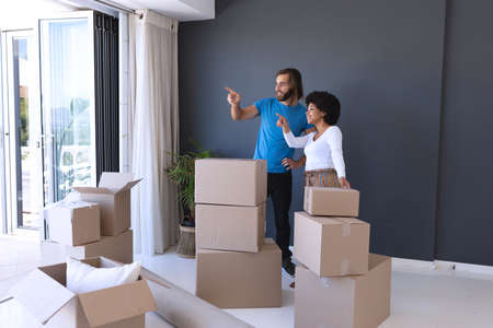 Happy diverse couple moving house smiling and pointing finger. staying at home in isolation during...