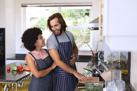 Happy diverse couple in kitchen preparing food and embracing. staying at home in isolation during...