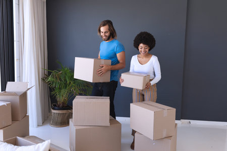 Happy diverse couple moving house smiling and holding cartons. staying at home in isolation during...