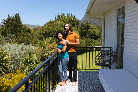 Happy diverse couple standing on balcony embracing and smiling. staying at home in isolation during...
