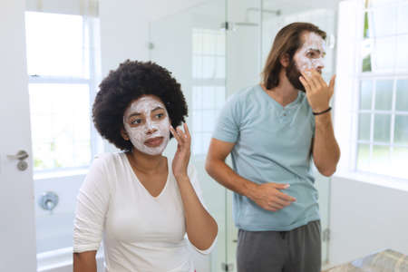 Diverse couple standing in bathroom wearing beauty masks. staying at home in isolation during...