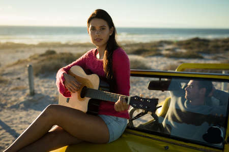 Caucasian woman sitting on beach buggy playing guitar, man sitting behind steering wheel. beach break on summer holiday road trip.の写真素材