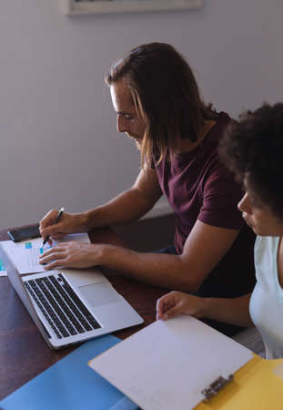 Diverse couple sitting at table working from home, using laptop and checking documents. staying at...