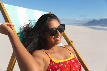 Smiling mixed race woman on beach holiday sitting in deckchair sunbathing. healthy outdoor leisure...